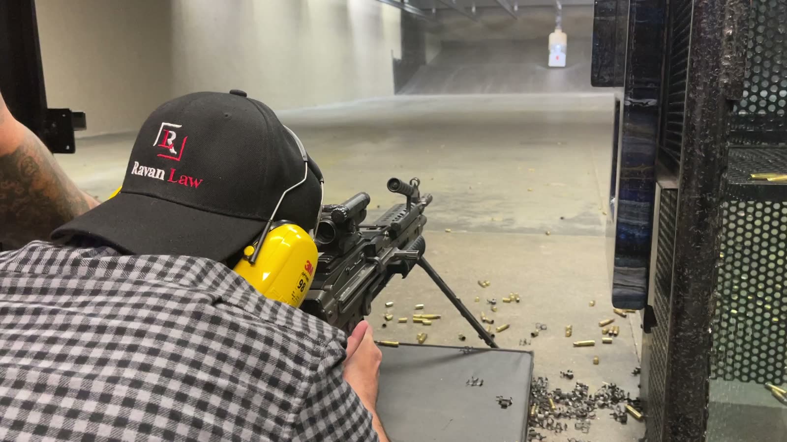 A shooter firing a belt-fed machine gun from a bench at an indoor range, spent brass on the floor.
