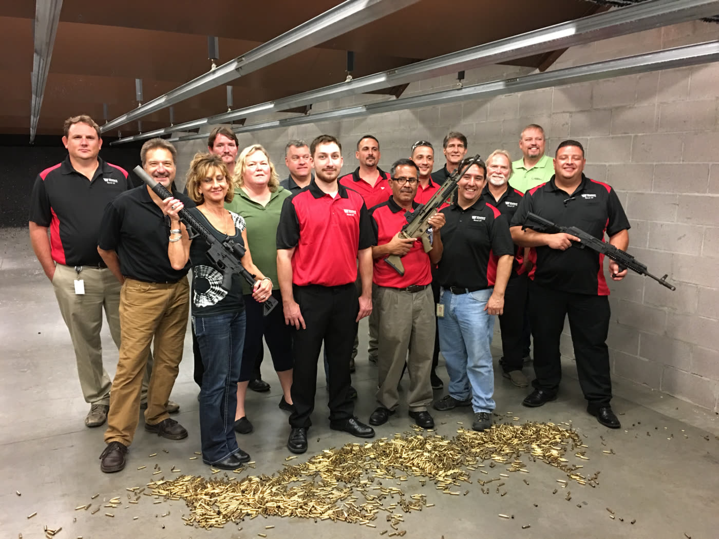 A large group of adults posing together with firearms at an indoor range, spent brass on the floor
